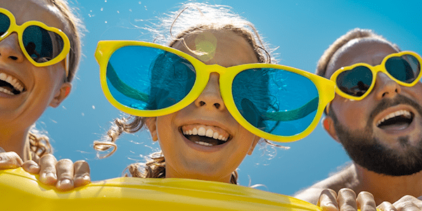 Elternpaar mit junger Tochter strampeln im großen gelben Schwimmring im Wasser und sind happy. Aller drei tragen lustige gelbe Sonnenbrillen bei strahlendem Sonnenschein.